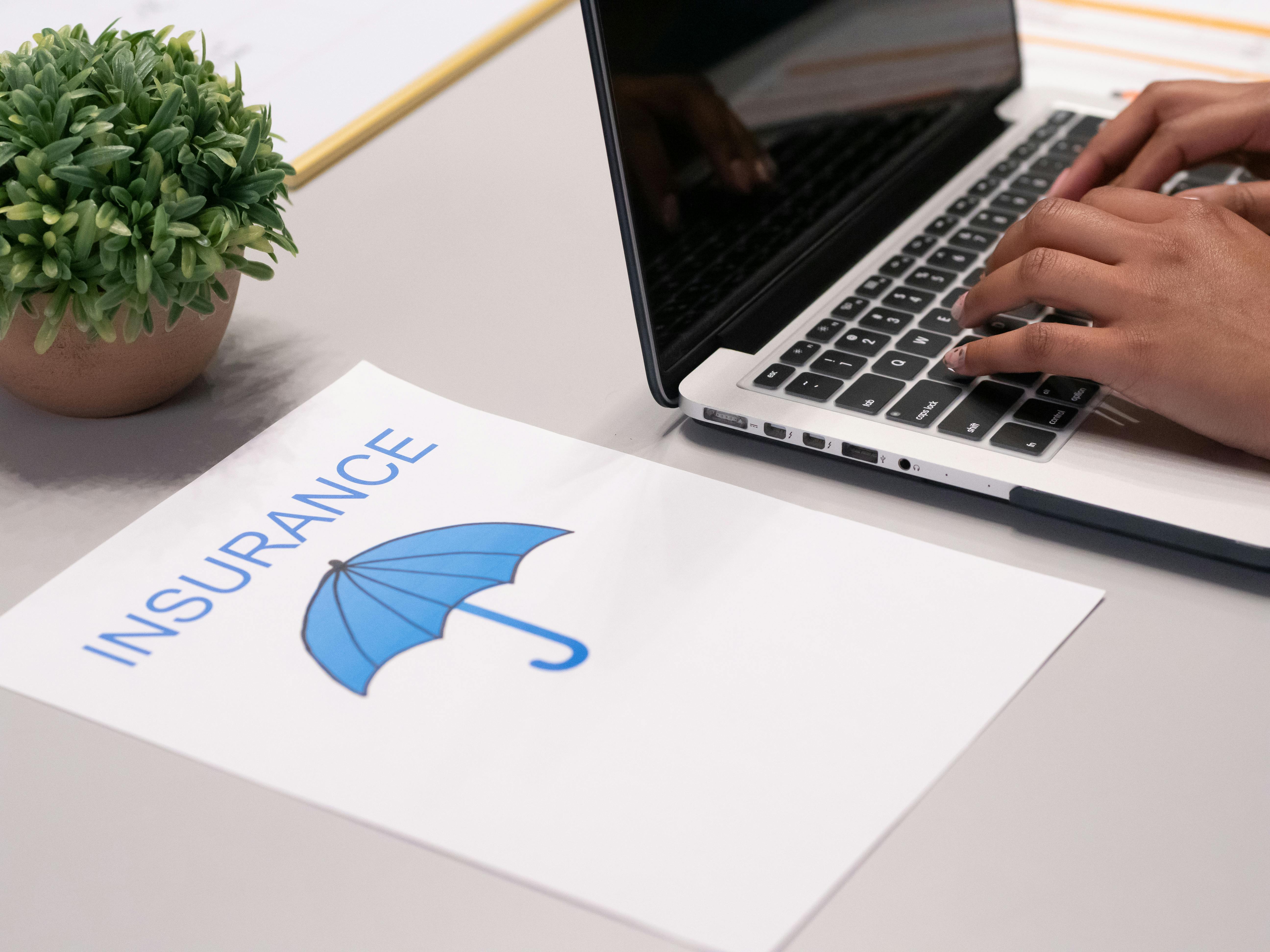 Photograph of someone typing on a laptop with a piece of paper next to them that has the word "Insurance" and an umbrella on it.