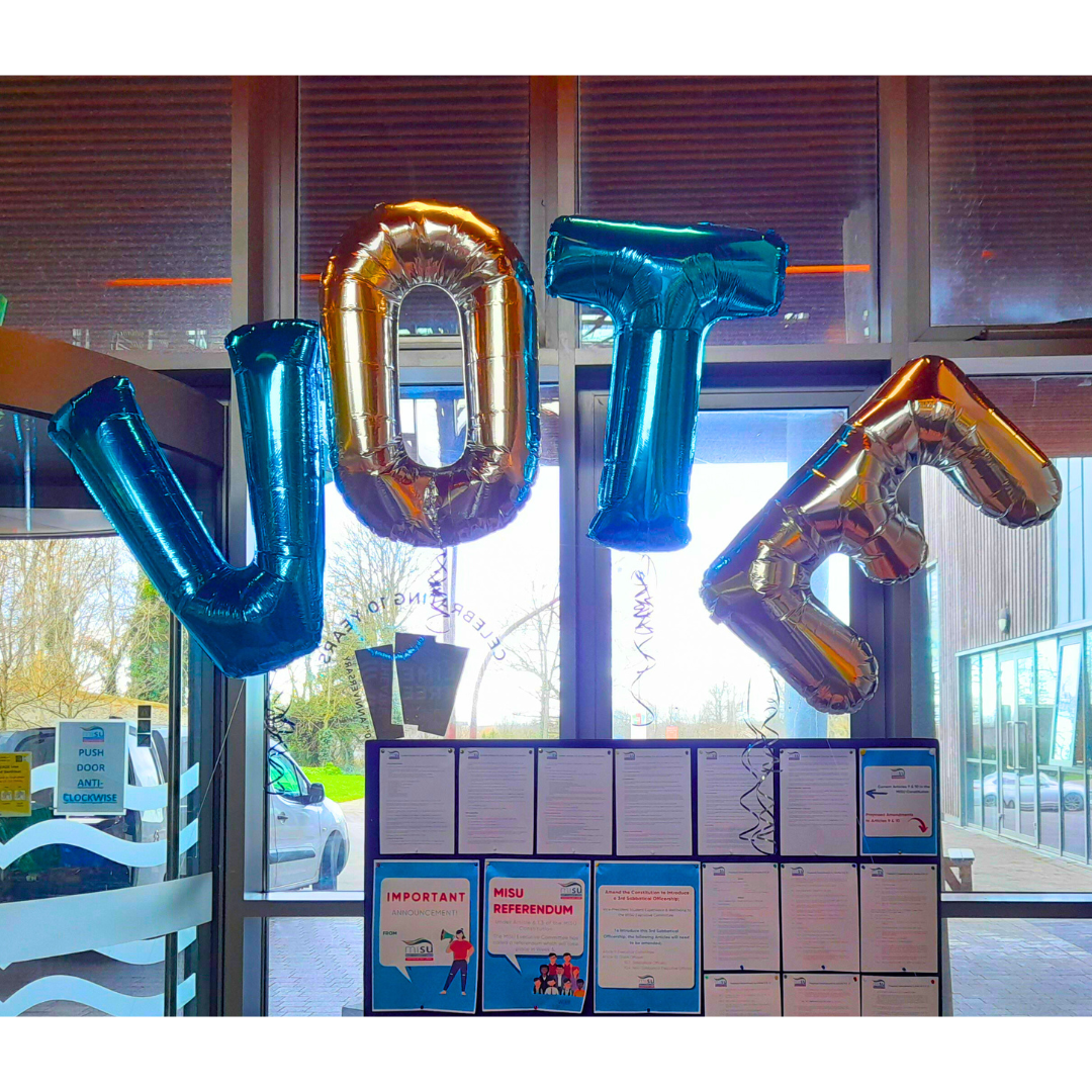 A photo of 4 balloons spelling out the word VOTE and a board with voting information displayed.